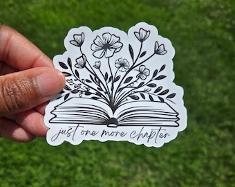 May include: A white sticker with a black floral design emerging from an open book. The text "just one more chapter" is written in a script font below the book.