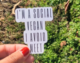 May include: White sticker with the text "I'M A SOCIAL VEGAN I AVOID MEET" in black, held against a background of green grass and leaves. The sticker has a unique shape and is held by a hand with red nail polish.