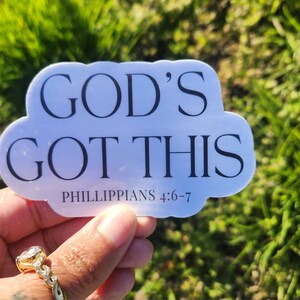 May include: A white sticker in a cloud shape with the words "GOD'S GOT THIS" in black, along with "PHILIPPIANS 4:6-7" in smaller text. The sticker is held in a hand with a gold ring, set against a blurred green background.