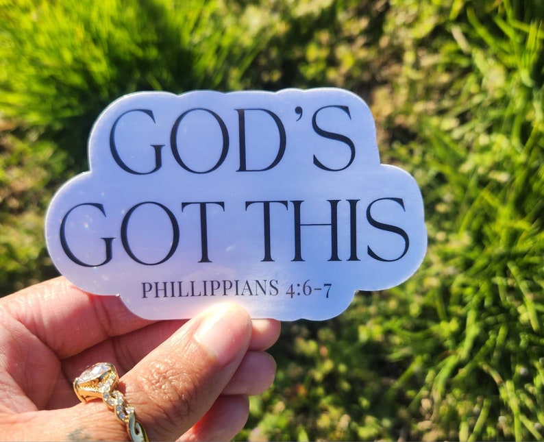May include: A white sticker in a cloud shape with the words "GOD'S GOT THIS" in black, along with "PHILIPPIANS 4:6-7" in smaller text. The sticker is held in a hand with a gold ring, set against a blurred green background.