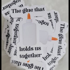 May include: White sticker with a glue bottle graphic and the text "The glue that holds us together." The glue bottle has an orange tip. The stickers are on a white, fluffy surface.