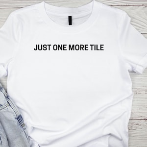May include: White t-shirt with the text "JUST ONE MORE TILE" in bold black letters. The shirt has short sleeves and a crew neck. Part of a pair of blue jeans is visible in the lower left corner.