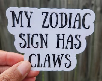 May include: A white sticker with black text that reads "MY ZODIAC SIGN HAS CLAWS". The sticker has a cloud-like shape and is held by a hand.