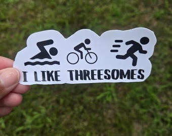 May include: A white sticker featuring black illustrations of a swimmer, cyclist, and runner, with the text "I LIKE THREESOMES" in black. The sticker is held against a backdrop of green grass.