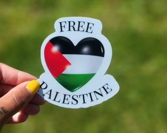 May include: A sticker featuring a heart-shaped design with the colors of the Palestinian flag: red, white, black, and green. The words "FREE PALESTINE" are printed on the sticker. The sticker is held against a blurred green background.