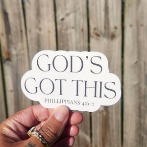 May include: A white sticker with the text "GOD'S GOT THIS" in a bold, serif font. Below the text is "PHILIPPIANS 4:6-7" in a smaller font. The sticker is held against a wooden fence.