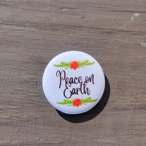 May include: A white button with the words "Peace on Earth" in cursive script, framed by green sprigs and red poinsettias. The button is round and has a glossy finish, perfect for holiday decorations.