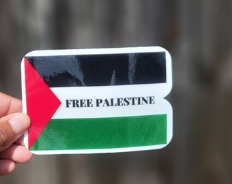 May include: A sticker featuring the Palestinian flag design with the words "FREE PALESTINE" in white. The flag design includes a red triangle, black, white, and green stripes. The sticker is held by a hand.