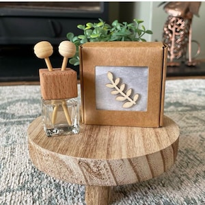 May include: A small glass bottle with a wooden top and two wooden sticks. The bottle is filled with a clear liquid. The bottle is sitting on a wooden stand. The bottle is next to a small cardboard box with a window that shows a wooden leaf design.