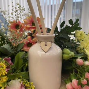 May include: A white ceramic diffuser bottle with a wooden heart charm. The bottle has three wooden diffuser sticks inserted into it. The bottle is surrounded by a bouquet of pink, yellow, and white flowers.