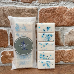 May include: Two wax melts, one in a white paper bag and one on a brick background. Both melts are white with blue glitter and the text "Laurel & Sage Candle Co."