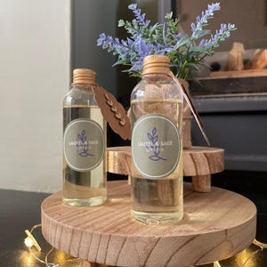 May include: Two clear glass bottles with gold lids and labels that say "Laurel & Sage Candle Co." on a wooden tiered stand. The bottles are filled with a light yellow liquid. A sprig of lavender is in the background.