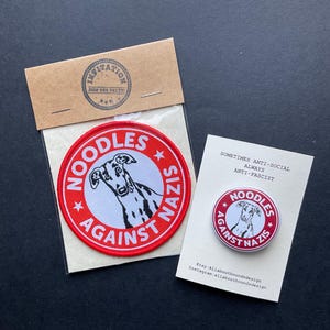 May include: A red and white patch and button featuring a dog illustration and the text "NOODLES AGAINST NAZIS". The patch is round and the button is on a white card with the text "Sometimes Anti-Social Always Anti-Fascist".