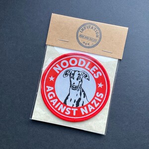 May include: A circular patch with the words "NOODLES AGAINST NAZIS" in red and white, surrounding a black and white dog illustration. The patch is attached to a brown card with the text "INVITATION JOIN OUR PARTY!"