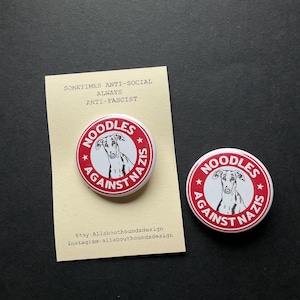 May include: Two round, red and white "Noodles Against Nazis" badges. Each badge features a greyhound illustration and text. A small card with text that reads "Sometimes Anti-Social Always Anti-Fascist" is also present.