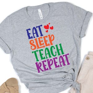 Puede incluir: Camiseta gris con el texto "Eat Sleep Teach Repeat" en colores arcoíris. Hay tres corazones rojos sobre la palabra "Eat".