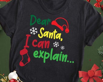 Può includere: T-shirt grigio scuro con la scritta "Dear Santa, I can explain..." in verde, giallo, rosso e bianco. Il design include un cappello di Babbo Natale, fiocchi di neve e una calza. Abbigliamento a tema natalizio.