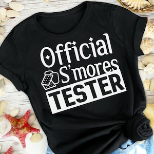 May include: Black t-shirt with white text that reads "Official S'mores Tester" and a graphic of a graham cracker and marshmallow.