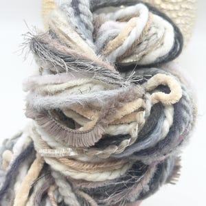 May include: A skein of art yarn featuring a blend of textures and fibers in shades of cream, beige, light gray, and charcoal. The yarn includes various strands such as fuzzy chenille, twisted cord, metallic ribbon, and soft wool.