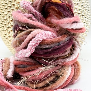May include: A bundle of assorted yarn in shades of pink, mauve, burgundy, and tan. The collection features various textures including fuzzy, braided, and metallic strands, wound together into a thick, decorative, and multi-textured fiber cluster.