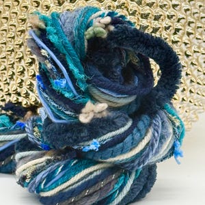 May include: A skein of art yarn featuring a variety of textures and shades of blue, including navy, teal, and light blue. The yarn incorporates metallic silver threads, fuzzy chenille, twisted cords, and soft white accents throughout the bundle.