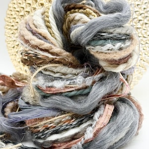 May include: A bundle of art yarn featuring a mix of textures and fibers in shades of grey, brown, cream, and copper. The yarn includes thick wool roving, twisted cord, metallic gold thread, and flat ribbon strands woven into a complex, layered coil.