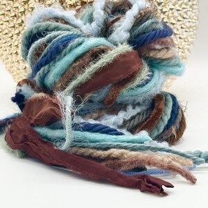 May include: A bundle of assorted yarn and ribbon in shades of light blue, teal, navy, brown, and tan. The collection features various textures including fuzzy, twisted, and smooth flat ribbon strands intertwined in a loose, layered arrangement.