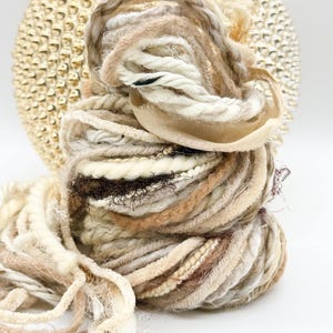 May include: A bundle of assorted yarn in neutral tones including cream, beige, tan, and dark brown. The collection features various textures such as thick twisted rope, fuzzy chenille, thin metallic strands, and smooth ribbon in a coiled arrangement.