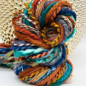 May include: A skein of art yarn featuring a blend of textures and fibers in shades of burnt orange, deep teal, navy blue, and mustard yellow. The yarn incorporates metallic gold ribbons, twisted cords, and fuzzy strands woven together.