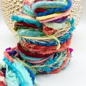 May include: A colorful, textured yarn bundle with a variety of threads and ribbons. The yarn features shades of blue, pink, red, yellow, and green. The bundle is tightly wound and has a decorative, handcrafted appearance.