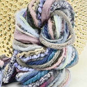May include: A skein of art yarn featuring a variety of textures and fibers in shades of blue, lavender, grey, and white. The yarn includes twisted cords, fuzzy mohair-like strands, and flat ribbon-like strips wound together in a dense, coiled bundle.