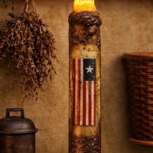 May include: A rustic, battery-operated candle with a distressed finish and a painted American flag design. The candle has a flickering, warm-toned LED flame. The candle is displayed on a wooden surface.
