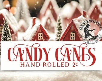 May include: A festive image featuring miniature red houses with snow-covered roofs and a banner that reads "CANDY CANES HAND ROLLED". The background includes blurred Christmas trees and falling snow, creating a winter holiday scene.