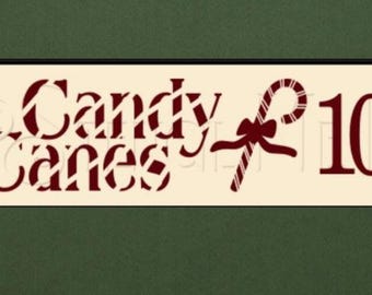 May include: A rectangular sign with the words "Candy Canes" in a decorative font, along with a candy cane graphic and the price "10¢". The sign is set against a cream background and framed in black. The background is a dark green with Christmas decorations.