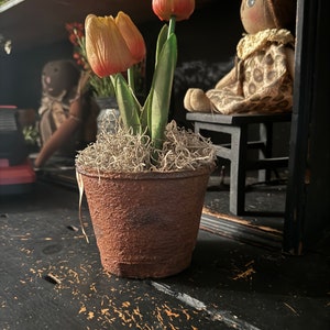 May include: A rustic, weathered metal pot filled with faux tulips. The tulips have yellow petals with red tips and green stems. The pot is filled with dried moss. A doll sits on a small black chair in the background.