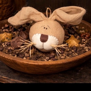 May include: A rustic wooden bowl filled with decorative elements, including a burlap bunny ornament with stitched details, star-shaped accents, and small brown stones. The bunny has long ears and a stitched face.