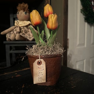 May include: A small, brown, textured pot with three yellow and red tulips. The pot has a tag that reads "Tulips". The tulips are surrounded by dried moss. A doll sits in the background.