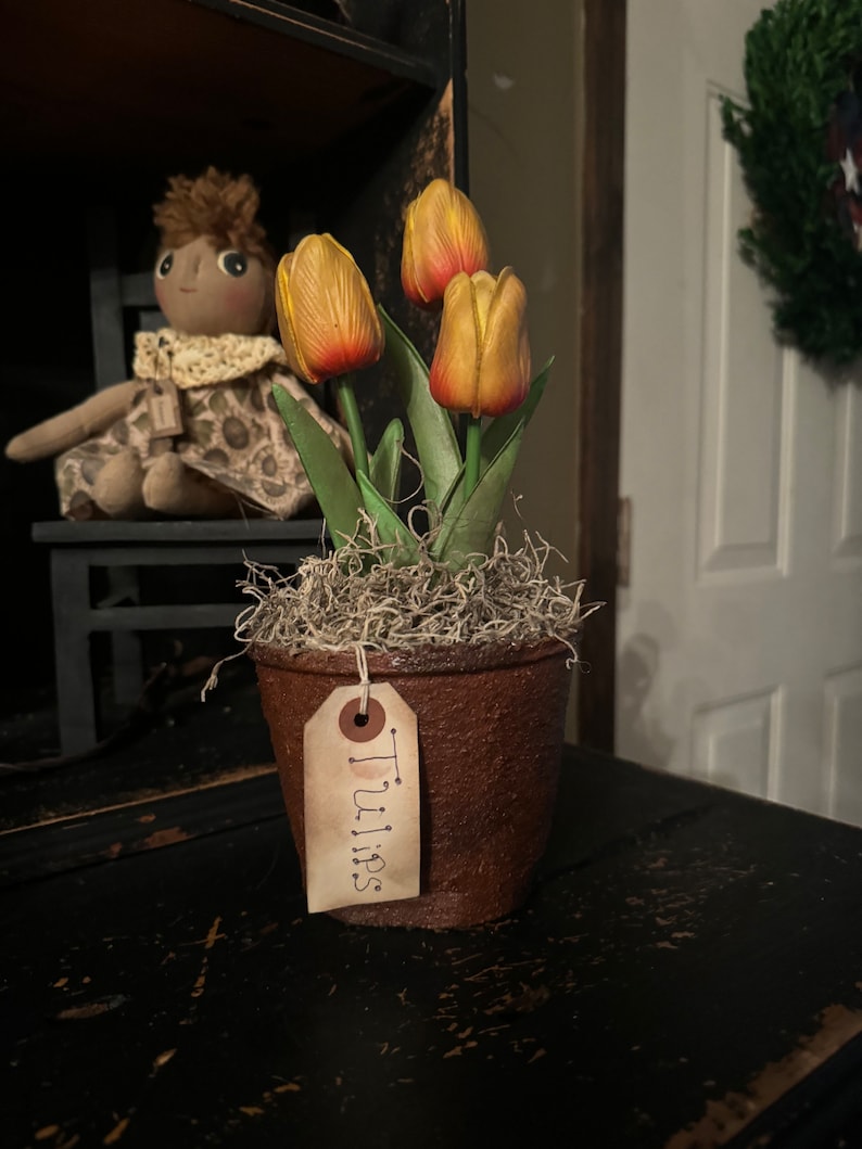 May include: A small, brown, textured pot with three yellow and red tulips. The pot has a tag that reads "Tulips". The tulips are surrounded by dried moss. A doll sits in the background.