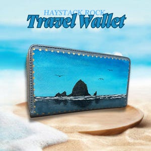 May include: A blue travel wallet with a painted image of Haystack Rock, a famous landmark in Oregon. The wallet has gold accents and a zipper closure.