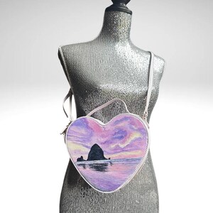 May include: A heart-shaped crossbody bag with a painted scene of a rocky island at sunset. The bag is pink and purple with a white trim.