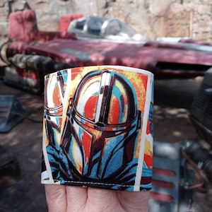 Puede incluir: Un brazalete de tela colorido con un estampado gráfico de un casco mandaloriano. El casco es azul, naranja y amarillo con una visera negra. La tela es blanca con un patrón amarillo, naranja y azul.