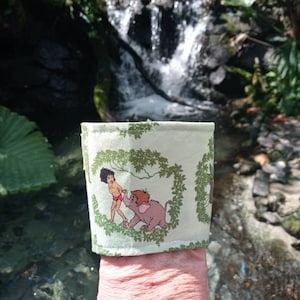 Könnte beinhalten: Ein grünes und weißes Stoffarmband mit einer Dschungelszene mit Mowgli und Baloo aus Disneys Dschungelbuch. Das Armband wird von einer Hand vor einem kleinen Wasserfall gehalten.