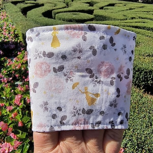 Pode incluir: Uma manga de tecido para copo com fundo branco e um padrão repetitivo de rosas rosa-pálido, folhas cinza-escuras e pequenas silhuetas amarelas de uma pessoa segurando um guarda-chuva. A manga possui bordas costuradas.