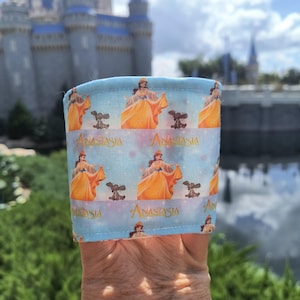 Pode incluir: Capa de copo de tecido azul claro com um padrão repetido de uma mulher em um vestido amarelo e um cachorro pequeno. A palavra "Anastasia" é impressa na capa. Um castelo é visível ao fundo.