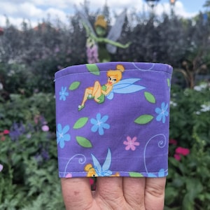 May include: A purple fabric drink sleeve with a cartoon fairy design. The sleeve is decorated with blue and pink flowers and green leaves. The fairy has blue wings and a yellow dress. The background is a garden.