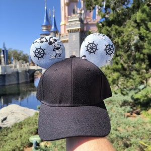 May include: Black baseball cap with blue Mickey Mouse ears featuring a nautical theme with white ship wheels and Mickey Mouse images.