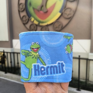 May include: A blue fabric wristband featuring a green Kermit the Frog graphic with the word "Kermit" in white. The wristband has a light blue interior and a repeating pattern of white radial lines.