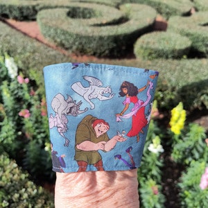 May include: A light blue fabric sleeve featuring characters from the animated film "The Hunchback of Notre Dame." The sleeve has illustrations of Quasimodo, Esmeralda, and gargoyles. The sleeve is wrapped around a cup.