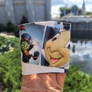 Peut inclure: Manchon de tasse à café en tissu blanc avec des illustrations de style bande dessinée de Jiminy Cricket de Pinocchio de Disney. Le manchon a un fond bleu et blanc avec le personnage dans diverses poses. Un château flou est en arrière-plan.