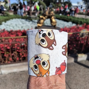 Puede incluir: Un brazalete de tela blanco con un patrón repetido de caras de ardilla de dibujos animados. Las ardillas son marrones y naranjas con ojos grandes y una nariz roja. El brazalete está doblado sobre la muñeca de una persona.